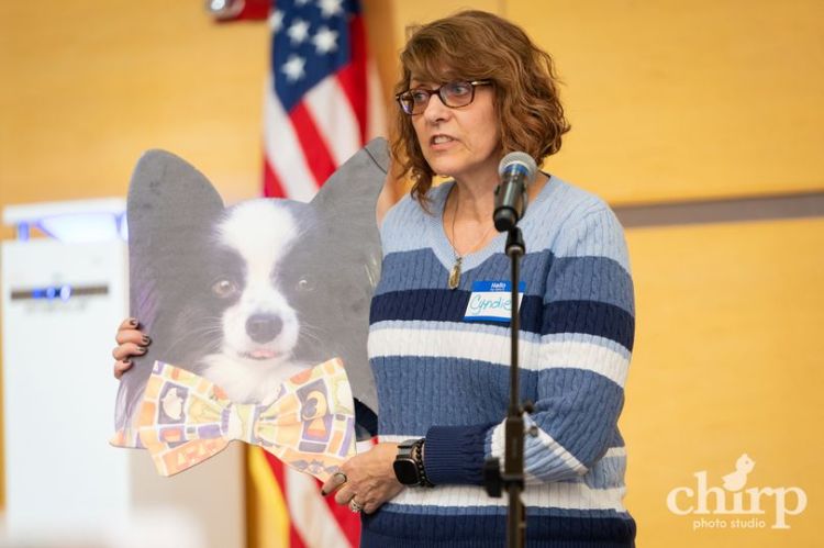 Photo of women at a podium holding a cardboard cut out of a black and white dog with erect ears.